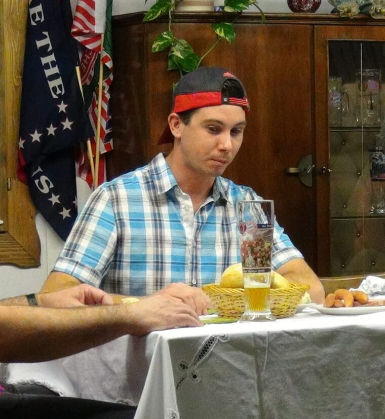 Junger Mann mit verkehrter Kappe und kariertem Hemd sitzt nachdenklich am gedeckten Tisch mit Weißbier, Brotkorb und Würstchen