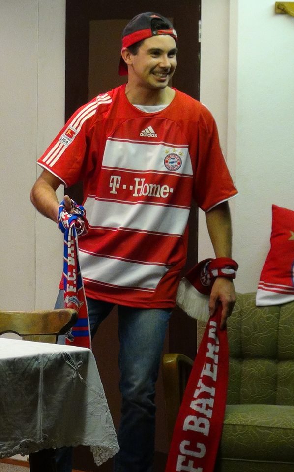 Junger Mann in rot-weißem FC-Bayern-Trikot und verkehrt aufgesetzter Kappe hält grinsend zwei Fanschals, hinten Sofa mit Bayern-Kissen