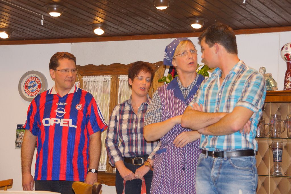 Mann im Bayern-Trikot, Frau in Karobluse, Frau mit Kopftuch und Kittelschürze und junger Mann mit verschränkten Armen in der Stube