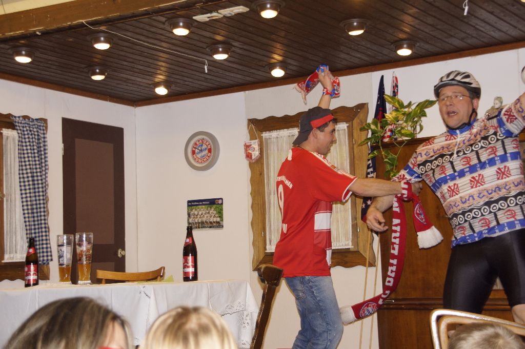 Junger Mann im Bayern-Trikot schwenkt jubelnd einen Fanschal, daneben Mann mit Radhelm und buntem Radtrikot, vorne Zuschauer