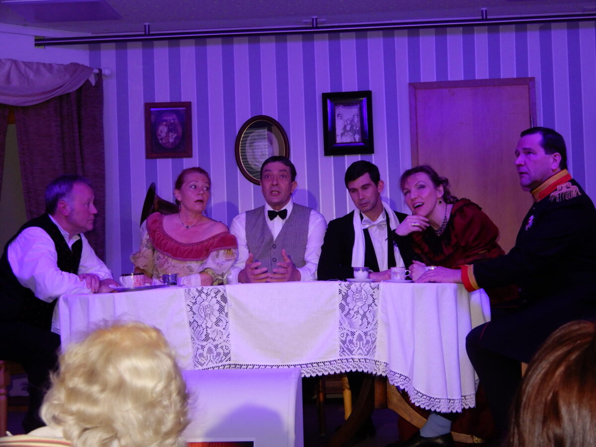 Sechs Darsteller in historischen Kostümen sitzen an einer Tafel mit Spitzendecke vor gestreifter Tapete, rechts ein Offizier