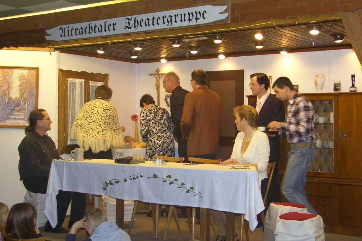 Aufbruch an der Festtafel: mehrere Gäste stehen auf und drängen nach hinten, eine Frau in Weiß und ein Mann bleiben sitzen