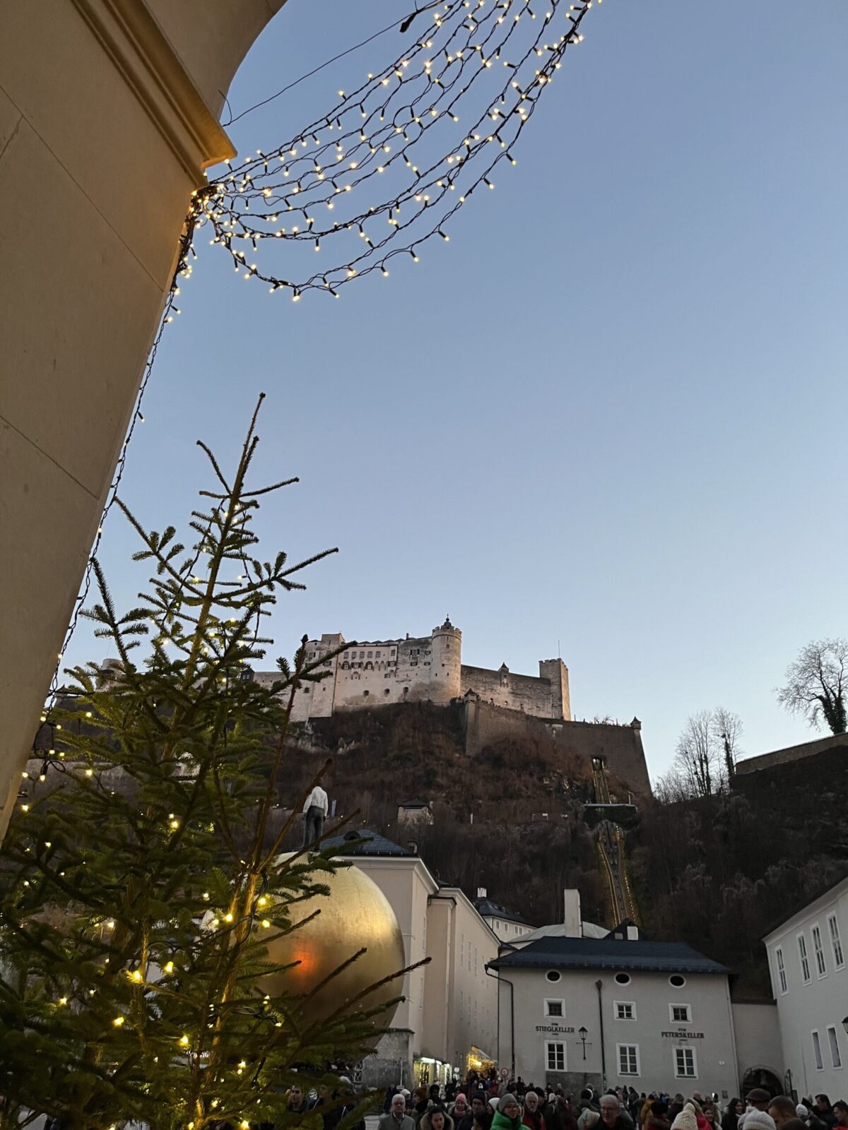 Festung Hohensalzburg in der Abenddämmerung über dem Kapitelplatz mit goldener Kugel, Lichterketten, Christbaum und vielen Menschen
