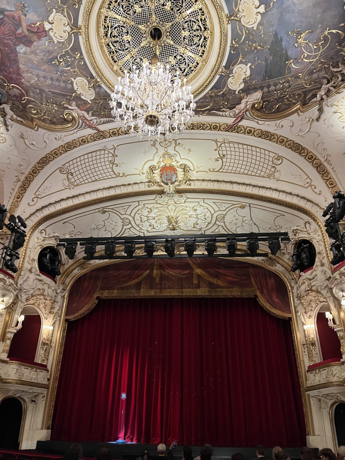 Zuschauerraum des Salzburger Landestheaters mit Kristalllüster, Deckengemälde, goldenem Stuck und geschlossenem rotem Samtvorhang, vorn Zuschauerköpfe