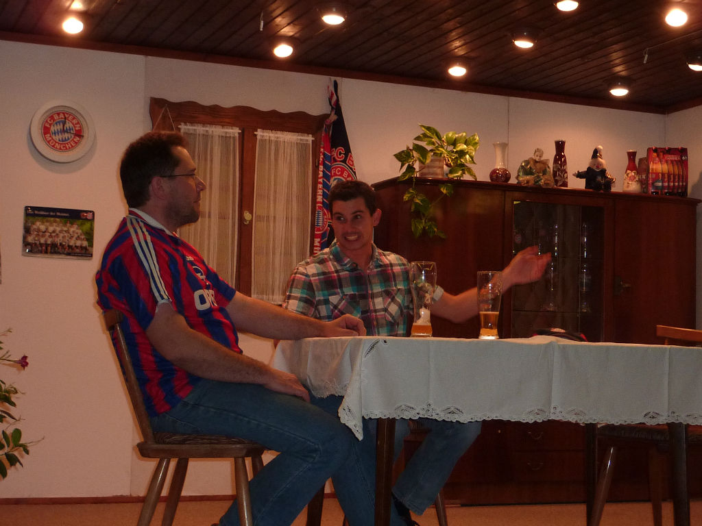 Mann im Bayern-Trikot und junger Mann im Karohemd sitzen mit Weißbiergläsern am Tisch, der Jüngere erzählt mit ausgestrecktem Arm