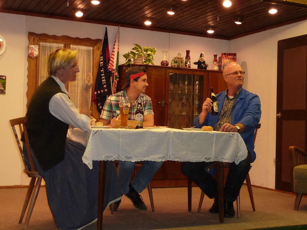 Frau mit Weste, junger Mann mit Kappe und Mann im blauen Kittel mit Bierflasche sitzen am Tisch und blicken lächelnd zur Seite