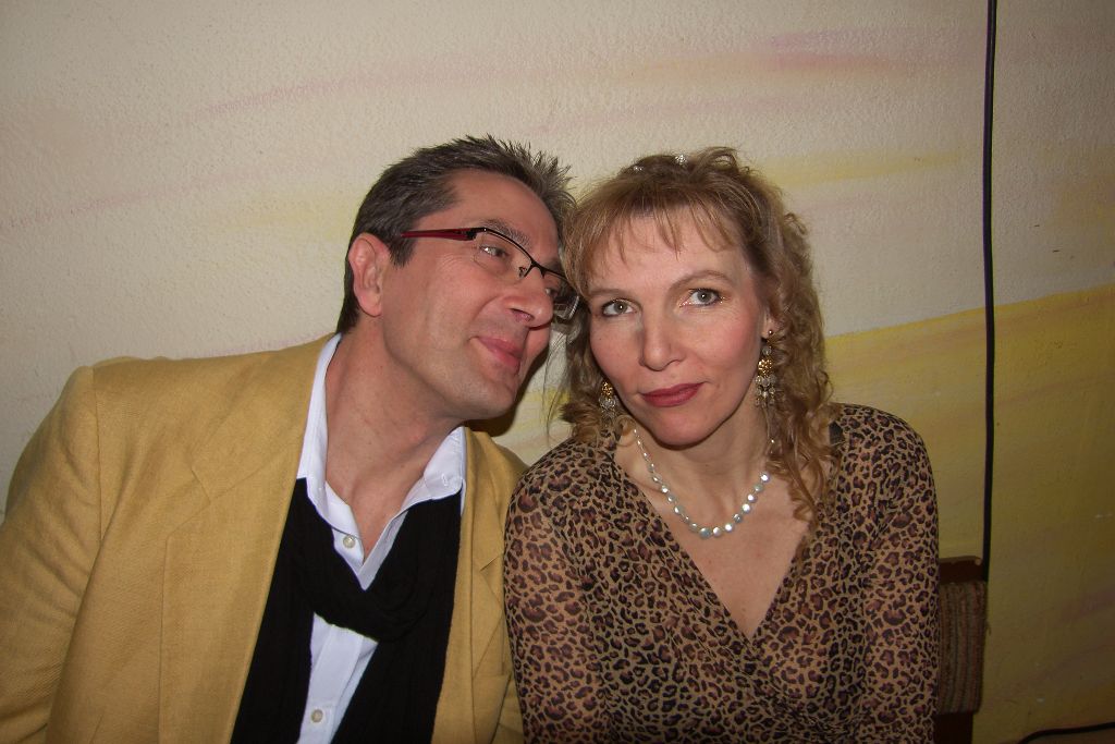 Mann im gelben Sakko mit schwarzem Schal und Frau im Leopardenkleid sitzen eng beieinander und lächeln in die Kamera