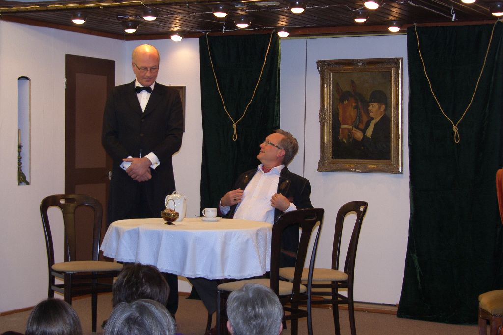Butler im Frack mit Fliege steht am runden Tisch, ein sitzender Herr im dunklen Sakko blickt lächelnd zu ihm auf; vorne Zuschauer