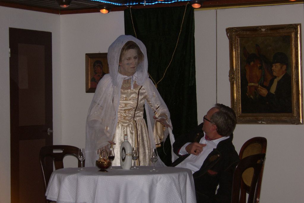 Die Ahnfrau als Gespenst in hellem Barockkleid und Schleier steht am Tisch, ein sitzender Mann im dunklen Sakko blickt zu ihr auf