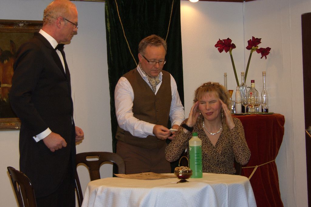Frau im Leopardenkleid hält sich am Tisch den Kopf, ein Mann in Weste reicht ihr einen Zettel, links steht der Butler im Frack