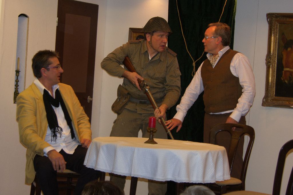 Mann in Soldatenuniform mit Stahlhelm und Gewehr wird von einem Mann in Weste gebremst, links sitzt ein Mann im gelben Sakko