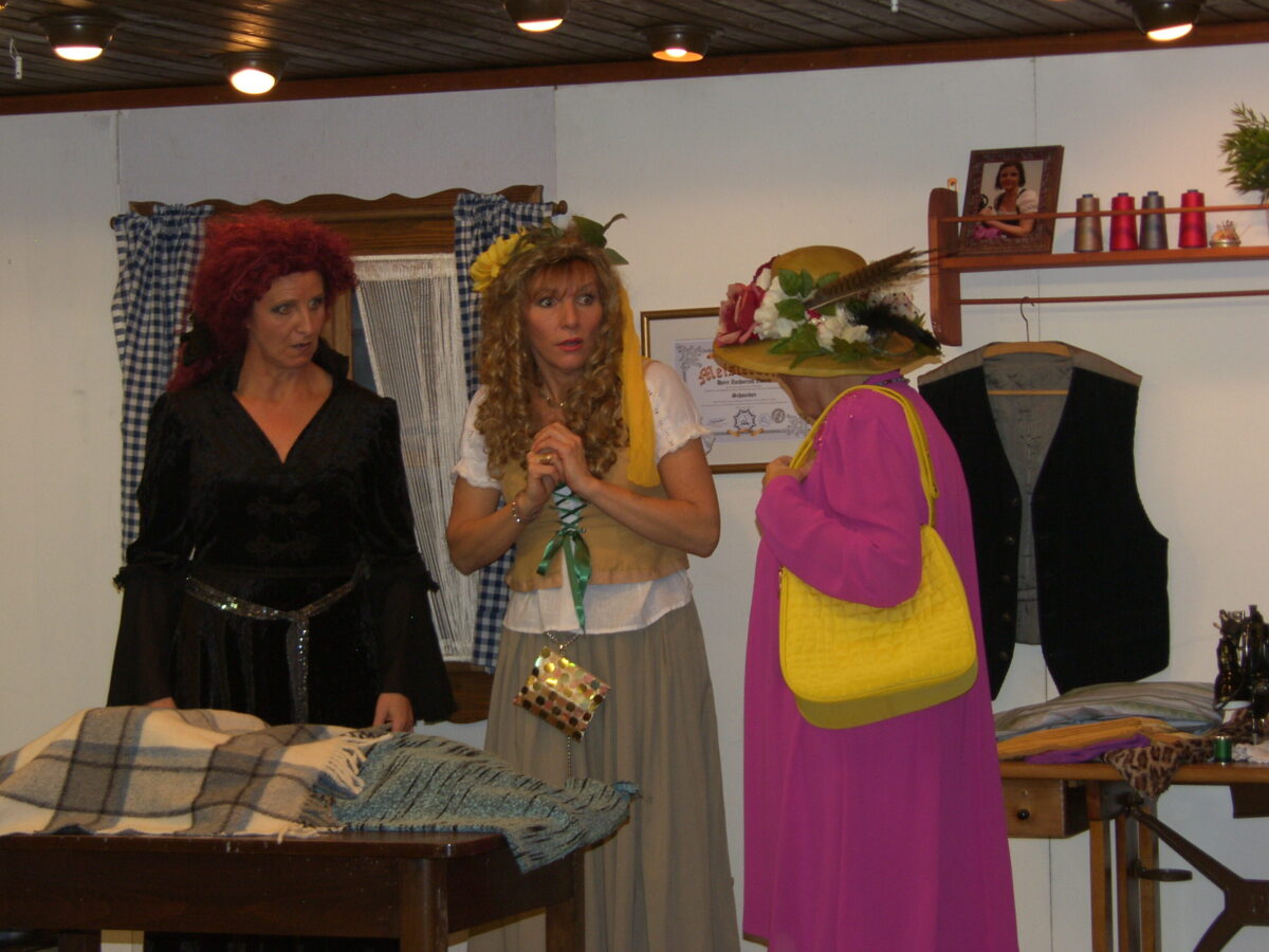 Rothaarige Hexe, blonde Hexe mit erschrockenem Blick und Frau mit Blumenhut und gelber Tasche stehen um einen Tisch mit Wolldecken