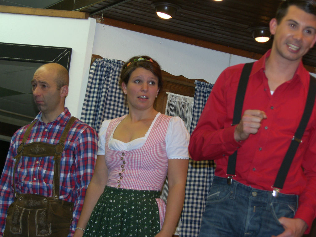 Mann in Lederhose, junge Frau im rosa Dirndl und lachender junger Mann in rotem Hemd mit Hosenträgern stehen nebeneinander