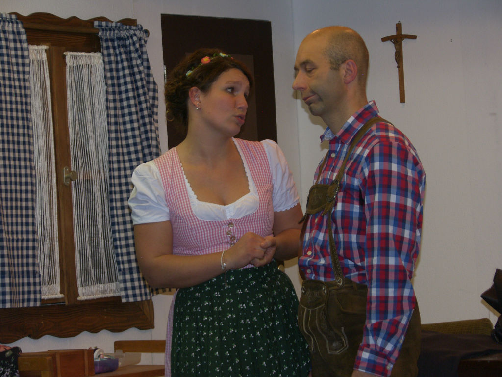 Junge Frau im rosa Dirndl mit Blumenhaarband redet eindringlich auf einen Mann in Lederhose und rot-blau kariertem Hemd ein