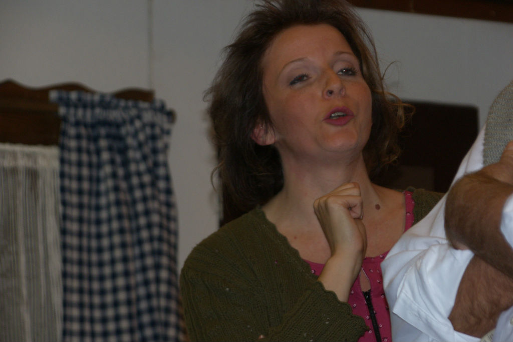 Nahaufnahme einer Frau mit zerzaustem Haar und grüner Strickjacke, die gestikulierend spricht, rechts angeschnitten ein Männerarm