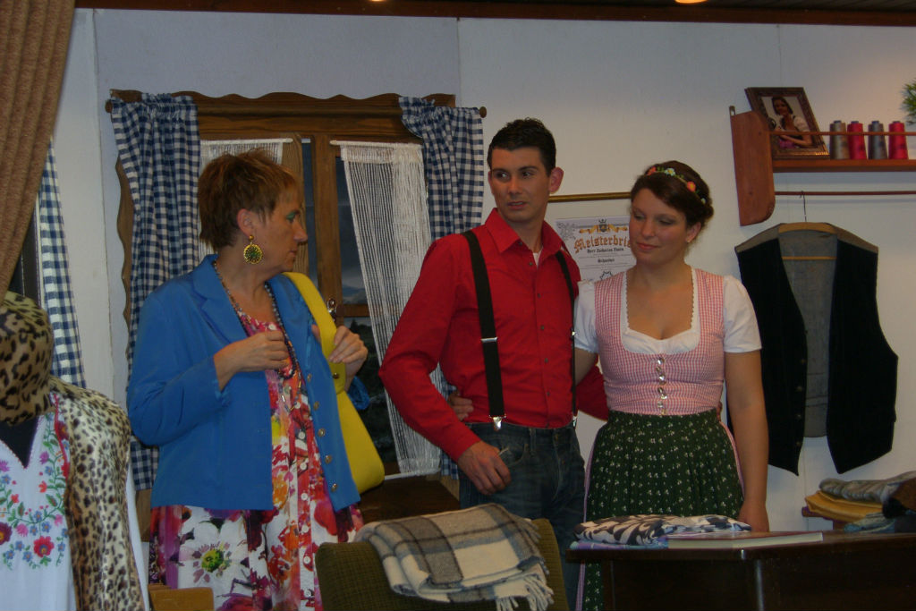 Frau in blauer Jacke und Blumenkleid mit gelber Tasche neben jungem Paar: Mann in rotem Hemd mit Hosenträgern, Frau im rosa Dirndl