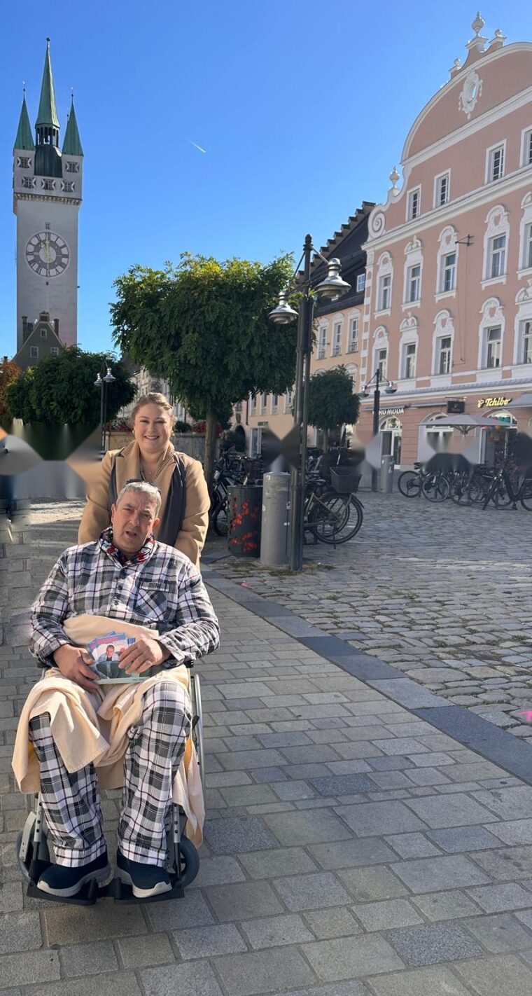 Werbeaktion am Straubinger Stadtplatz: Mann im karierten Schlafanzug sitzt mit Decke und Flyern im Rollstuhl, eine Frau schiebt ihn