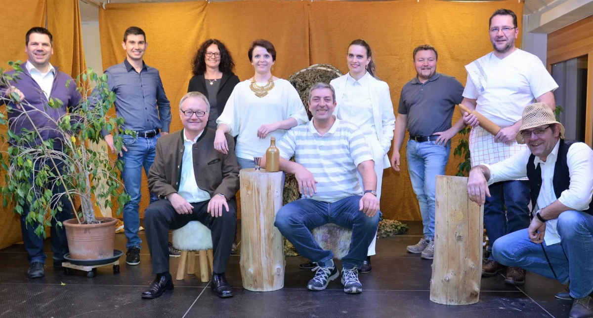 Elf Mitglieder der Aitrachtaler Theatergruppe vor einem goldgelben Bühnenvorhang, einige sitzen auf Baumstümpfen