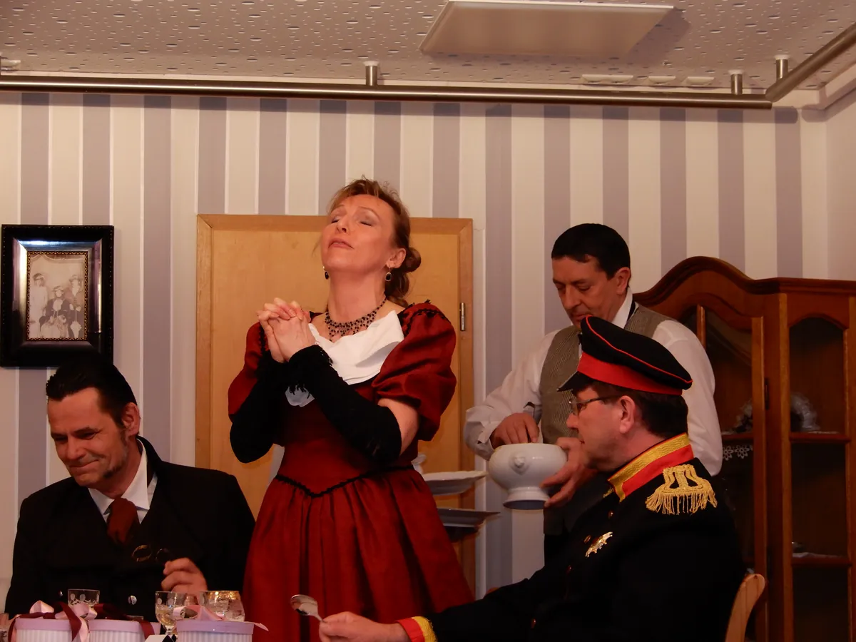 Szene mit einer Frau im roten Kleid, einem Mann in Uniform und Gästen an der Tafel