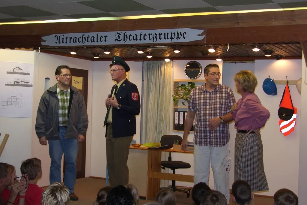 Bühnenbild eines Büros mit Aktenordnern, vier Darsteller und ein Polizist, darüber das Banner der Aitrachtaler Theatergruppe