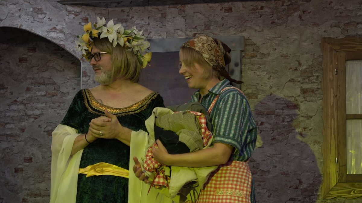 Figur im grünen Samtkleid mit Blumenkranz im Haar neben lachender Magd mit Kopftuch, karierter Schürze und Wäschebündel