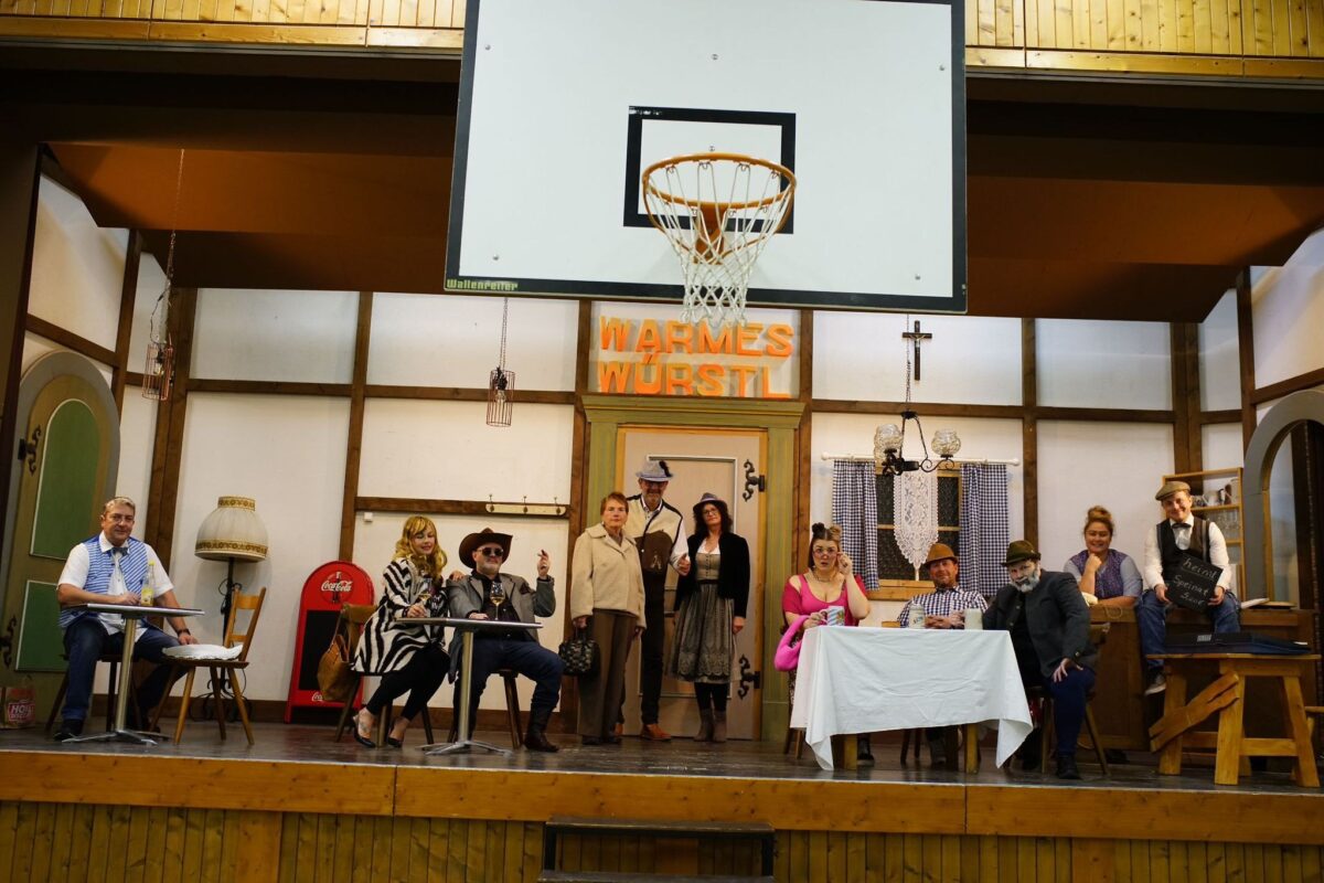 Ensemble in Kostümen im Kneipen-Bühnenbild „Warmes Würstl“, darüber der Basketballkorb der Mehrzweckhalle Aiterhofen