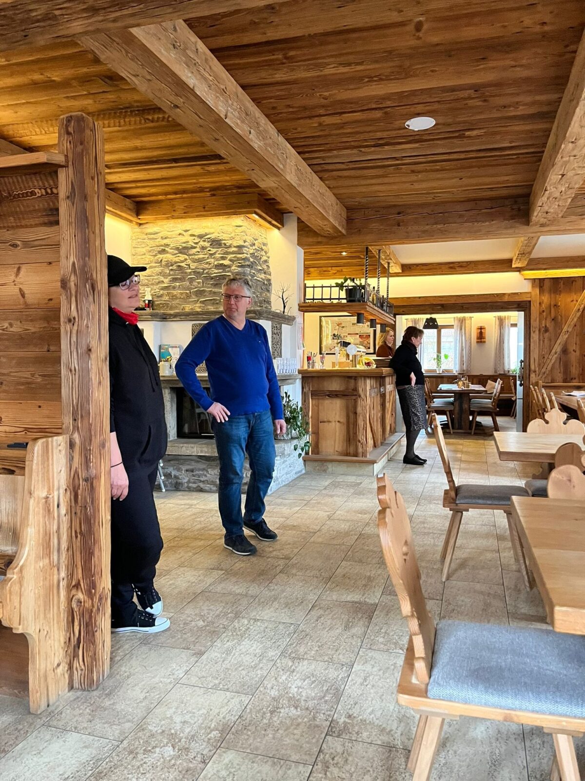 Probe in der Gaststube: Darstellerin mit Kappe an einem Holzpfosten, Mann im blauen Pullover, hinten Theke und Kamin