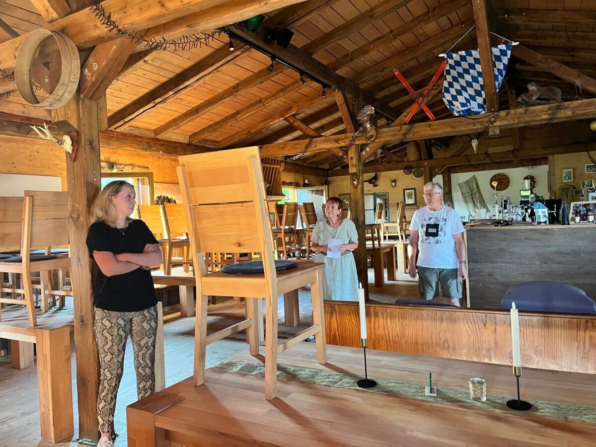 Probe in einer rustikalen Hütte: Frau lehnt an einem Holzbalken, zwei Mitspieler mit Textblatt, Stühle auf den Tischen