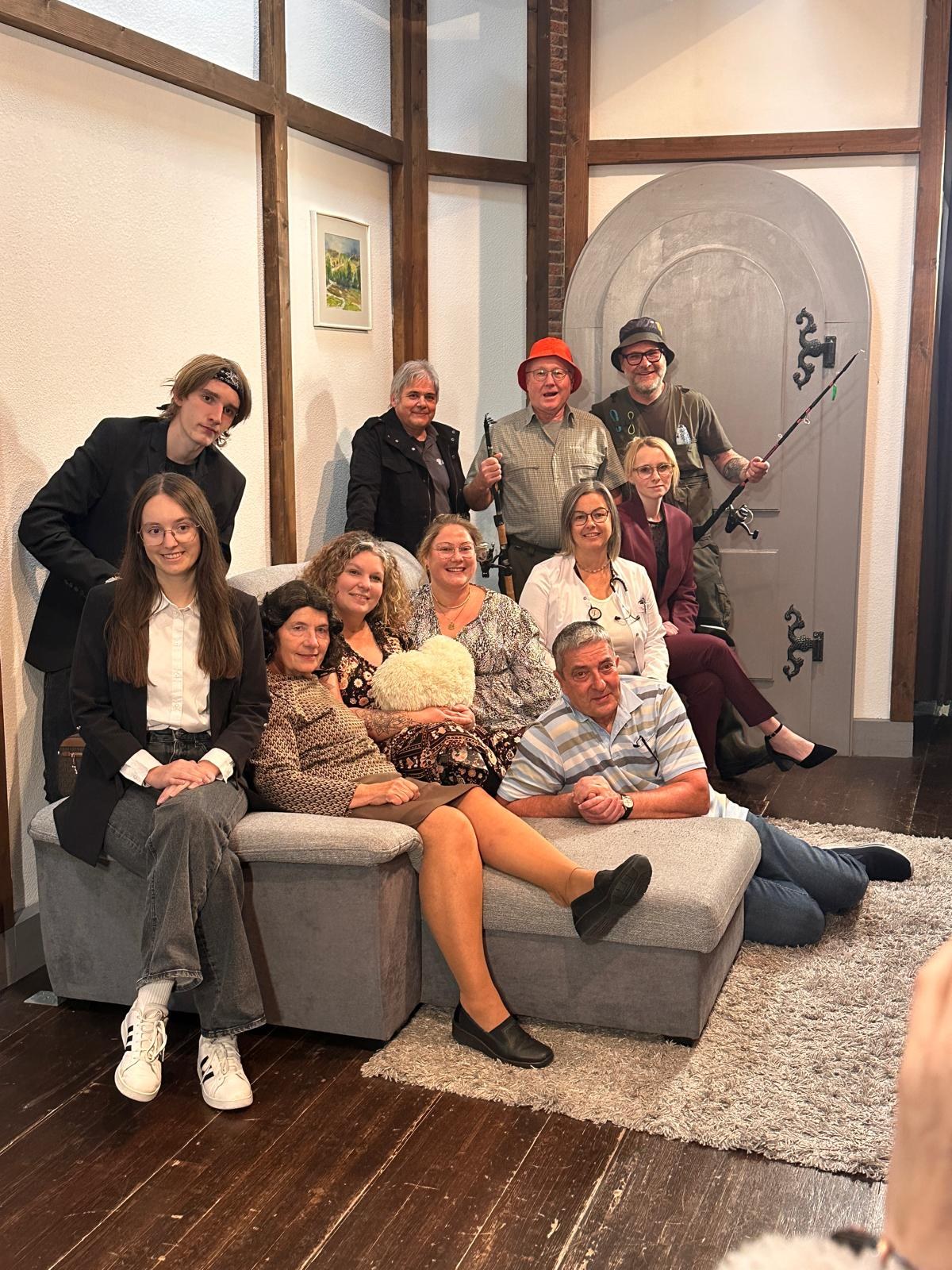 Ensemble von „Männergrippe“ im Bühnenbild: elf Darstellerinnen und Darsteller auf und um ein graues Sofa vor einer grauen Rundbogentür, darunter zwei Angler mit Hut und Angelrute und eine Ärztin im weißen Kittel
