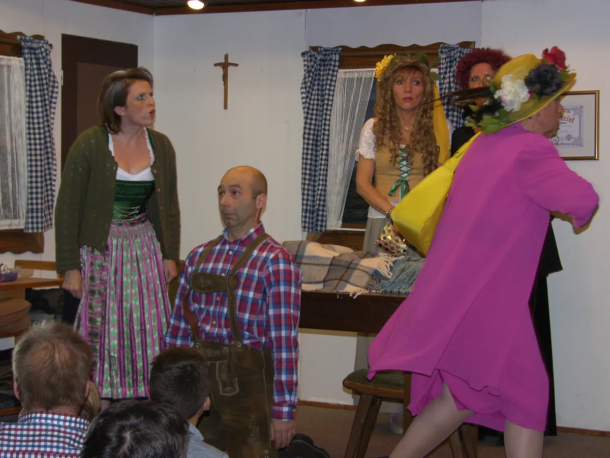 Zwei Wetterhexen in bunten Kostümen, ein Mann in Lederhose und eine Frau im Dirndl in einer Stube