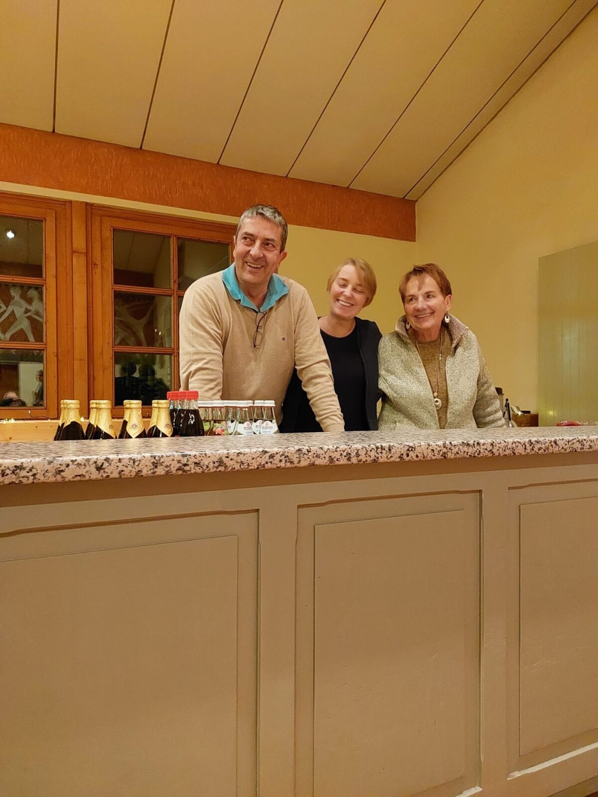 Ein Mann und zwei Frauen stehen lächelnd hinter einer Theke mit Getränkeflaschen