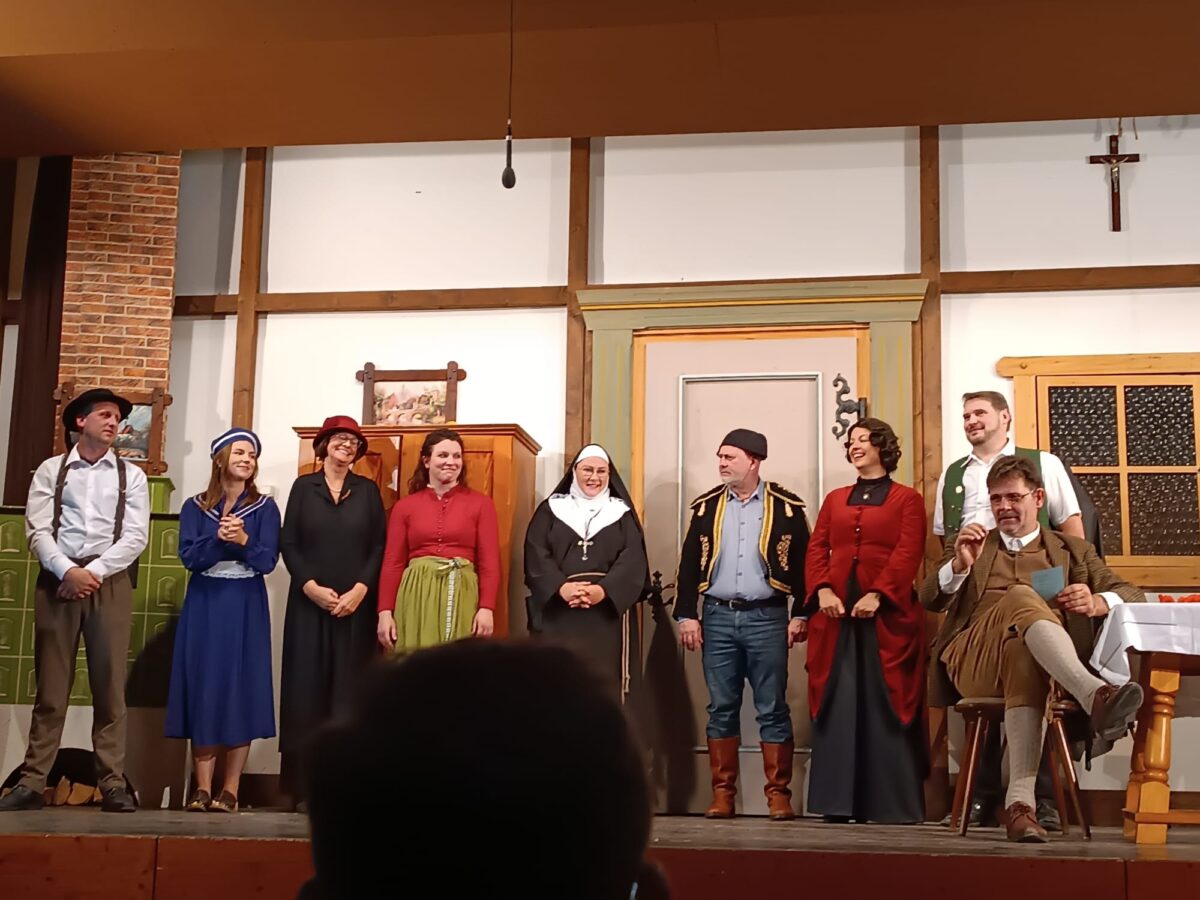 Ensemble in Kostümen steht in einer Reihe vor der Bauernstuben-Kulisse, darunter Nonne, Matrosenkleid und Trachtenweste