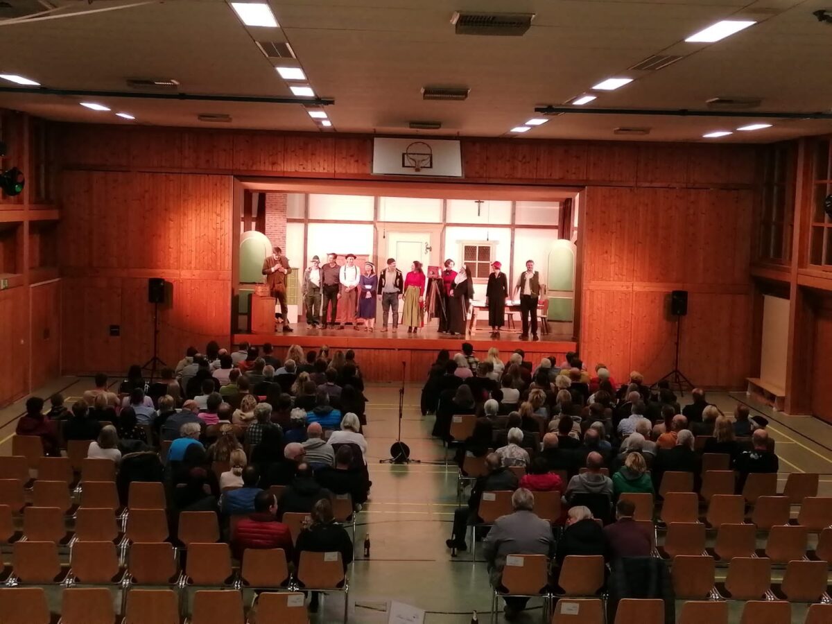 Blick über das volle Publikum der Mehrzweckhalle Aiterhofen auf das Ensemble beim Schlussapplaus auf der Bühne