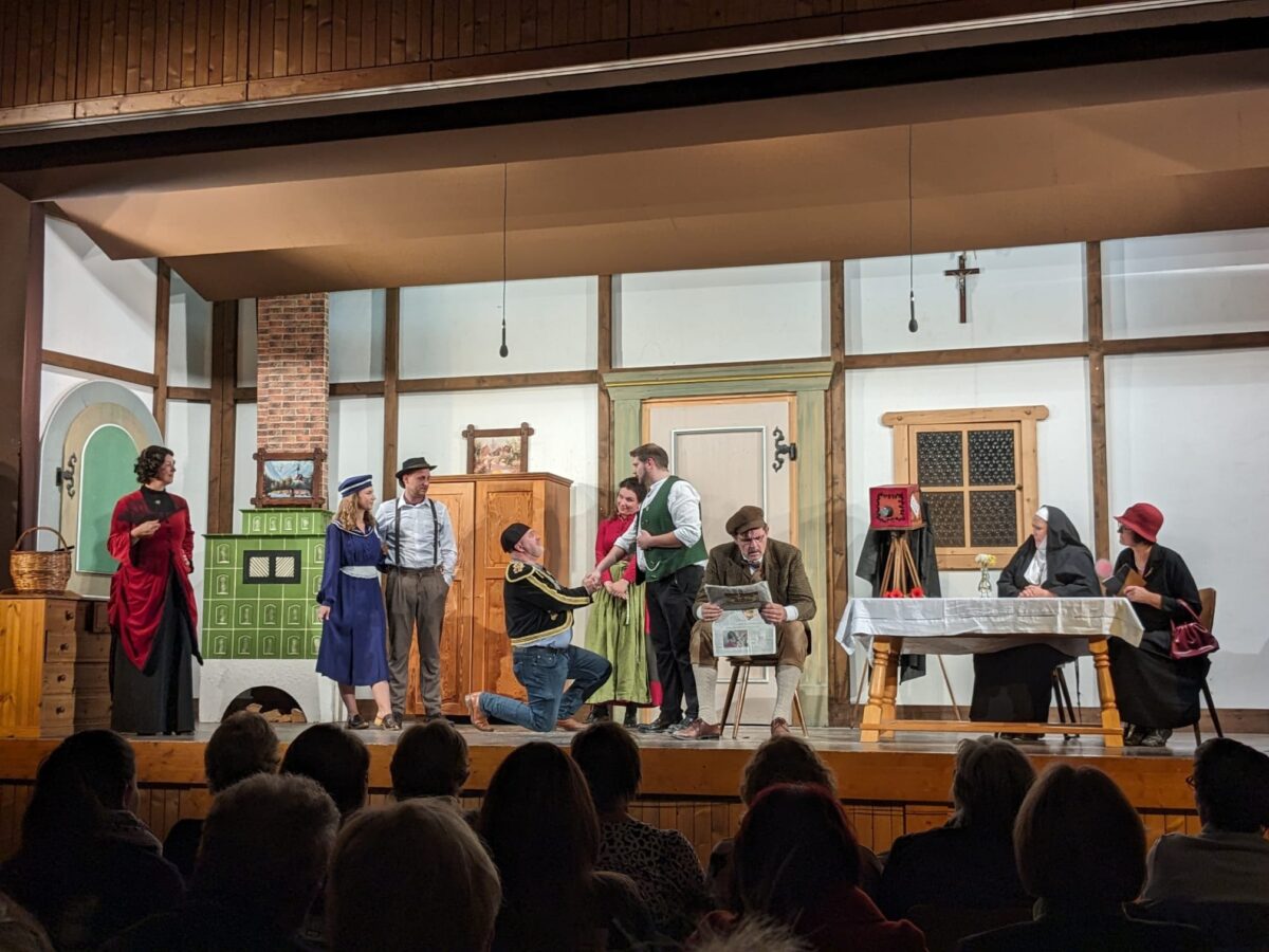 Szene in der Bauernstube: Mann kniet und hält die Hand einer Frau im Dirndl, andere schauen zu, Nonne und Dame am Tisch