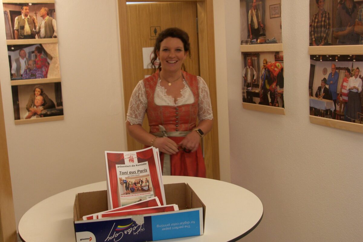 Frau im roten Dirndl hinter einem Tisch mit Programmheften zu „Toni aus Paris“, an der Wand Fotos früherer Aufführungen