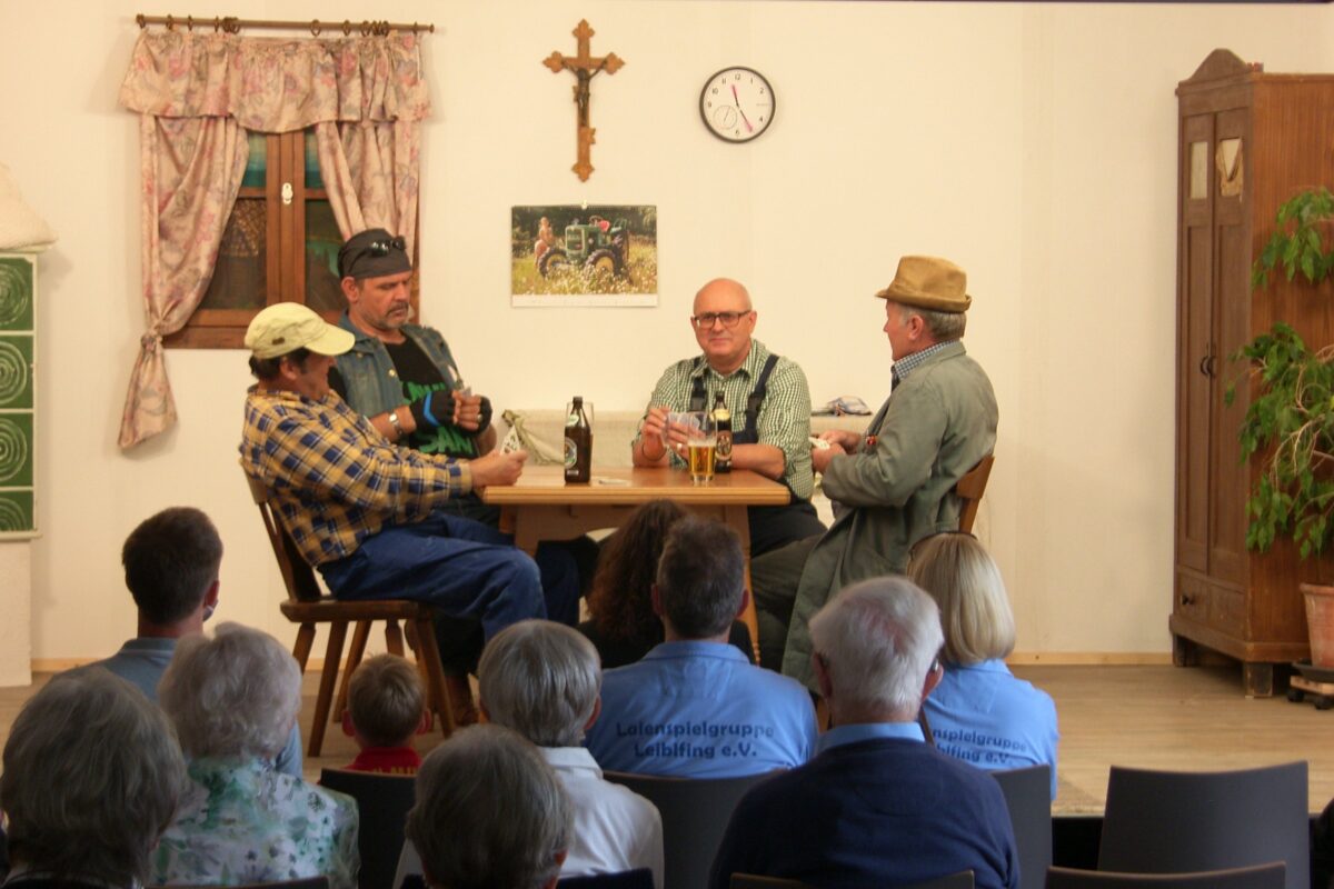 Vier Männer spielen am Stubentisch mit Bierflaschen Karten; Bauernstube mit Kruzifix, Uhr und Schrank, vorn das Publikum