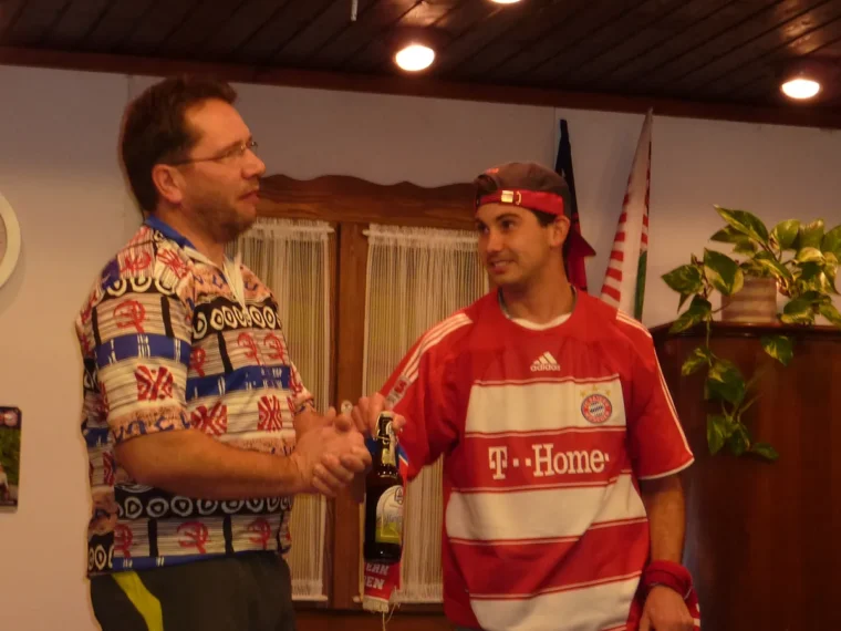 Ein junger Mann im FC-Bayern-Trikot mit Stirnband schüttelt einem Mann im bunten Hemd die Hand